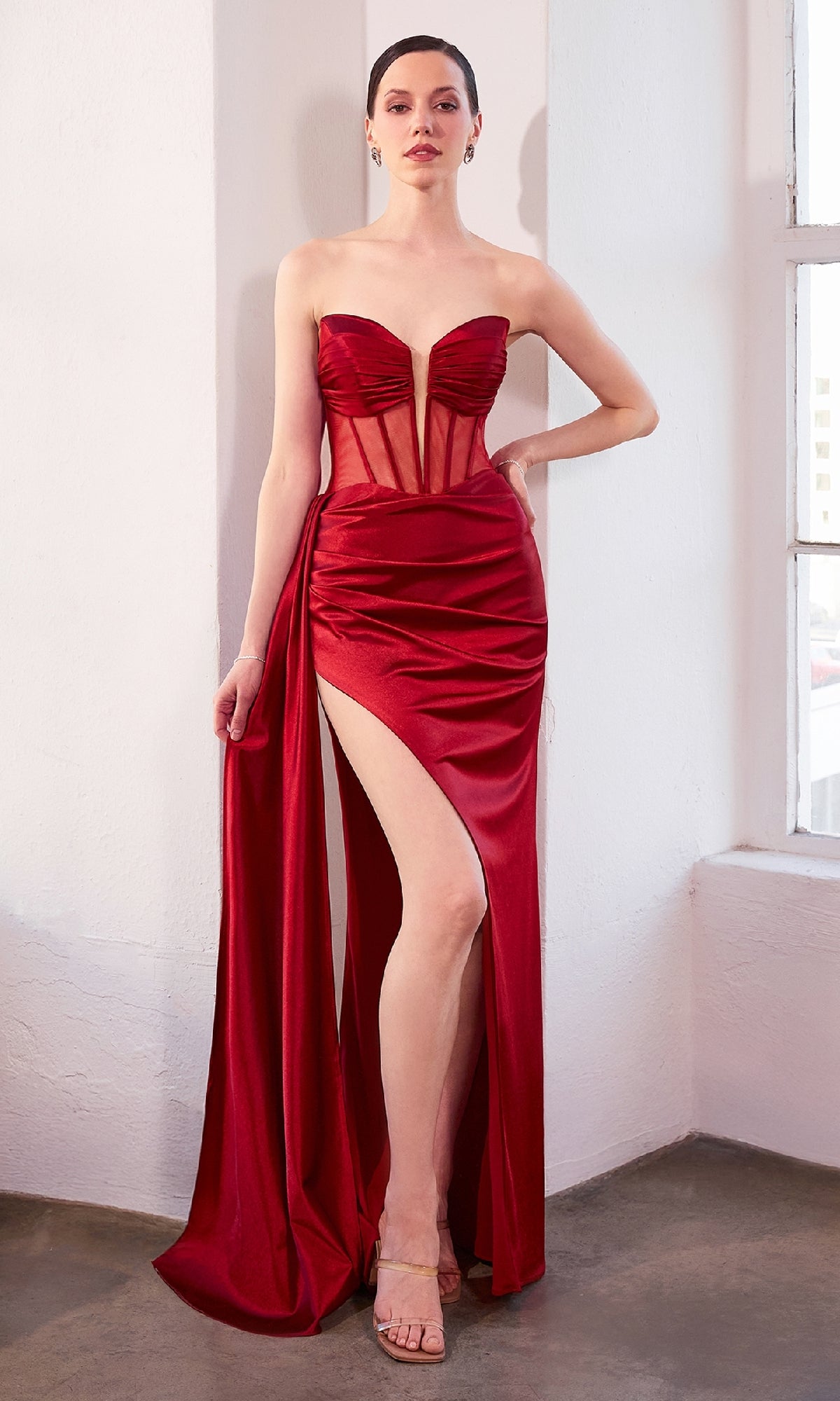 A woman in the Strapless Deep V-Neck Long Prom Dress: Amoris T811, featuring red satin with a sheer panel and high thigh slit, stands indoors against a white wall.